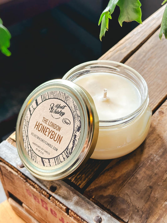 A small straight-sided jar candle with a gold lid, labeled 'The London Honeybun'. The candle is shown both with the lid on and off, revealing the candle wax inside.